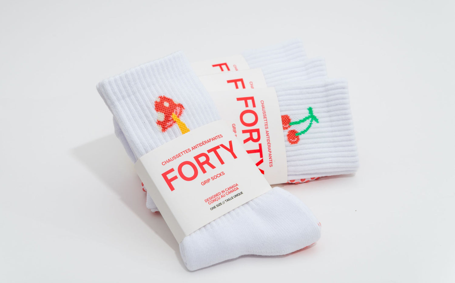 A white pair of FORTY grip socks with a playful red and yellow mushroom lean against a pair of cherry FORTY pilates socks.
